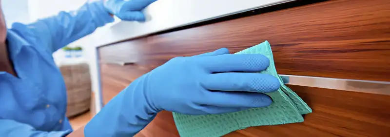 Cleaner wearing blue gloves wiping a wooden cabinet drawer with a microfiber cloth for dust and grime removal, Pasadena, CA.
